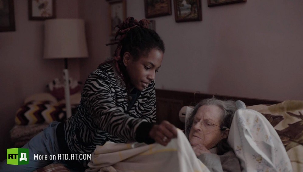 Young African American woman holding up sheets to tuck in an elderly White woman in bed. Still taken from RTD Documentary film Black Lives 6. Illusion about single-parent families and teenage parenthood in Black America.