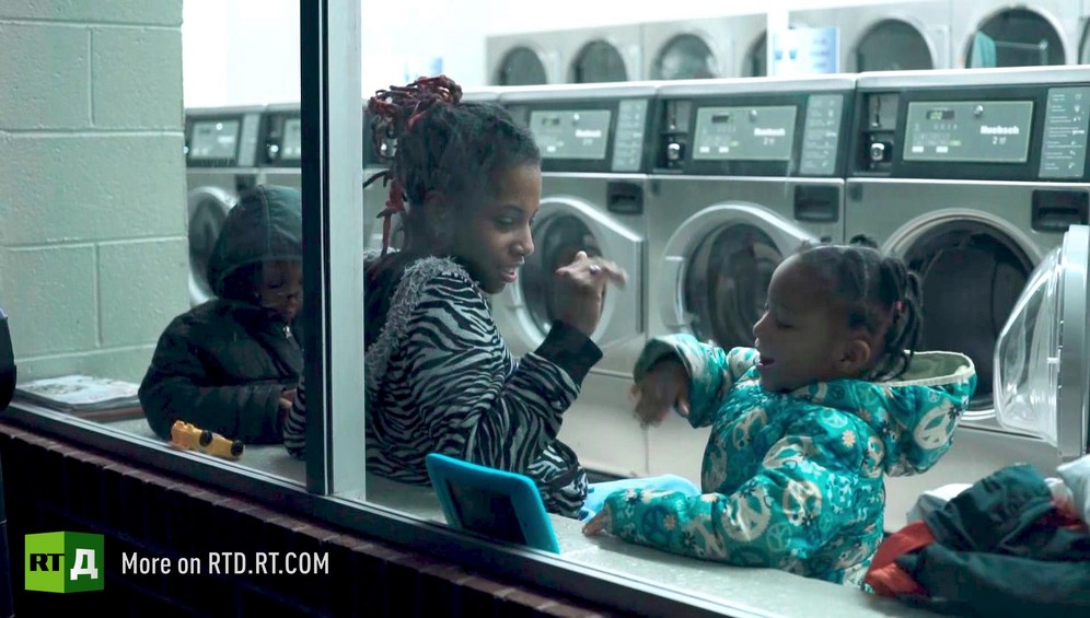 African American mother interacting with her daughter, who is playing on a tablet, while her son sits next to her in a laundromat. Still taken from RTD Documentary film Black Lives 6. Illusion about single-parent families and teenage parenthood in Black America.