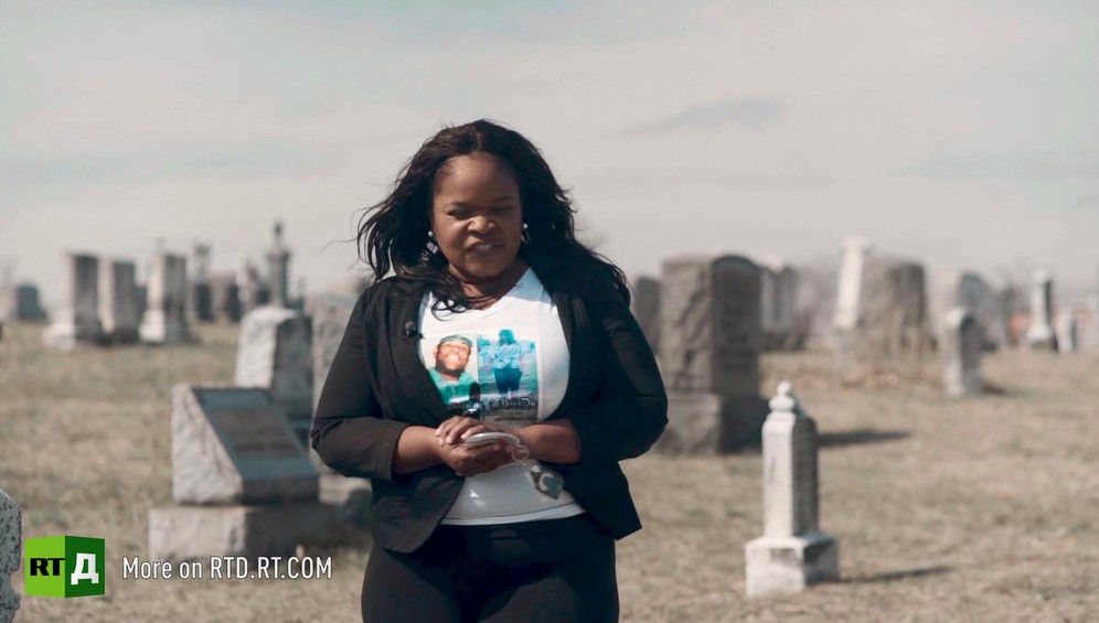 African American woman wearing a t-shirt with pictures of sons who were shot, standing in front of headstones in a cemetery in the sun. Still taken from RTD Documentary film Black Lives 6. Illusion about single-parent families and teenage parenthood in Black America.