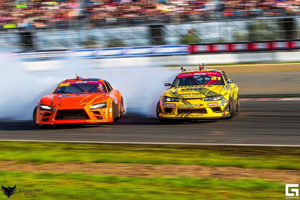 An orange and a red car emit smoke as they skid side by side around a corner of the track during Russian Drift Series competition in the  2018 drift racing championship
