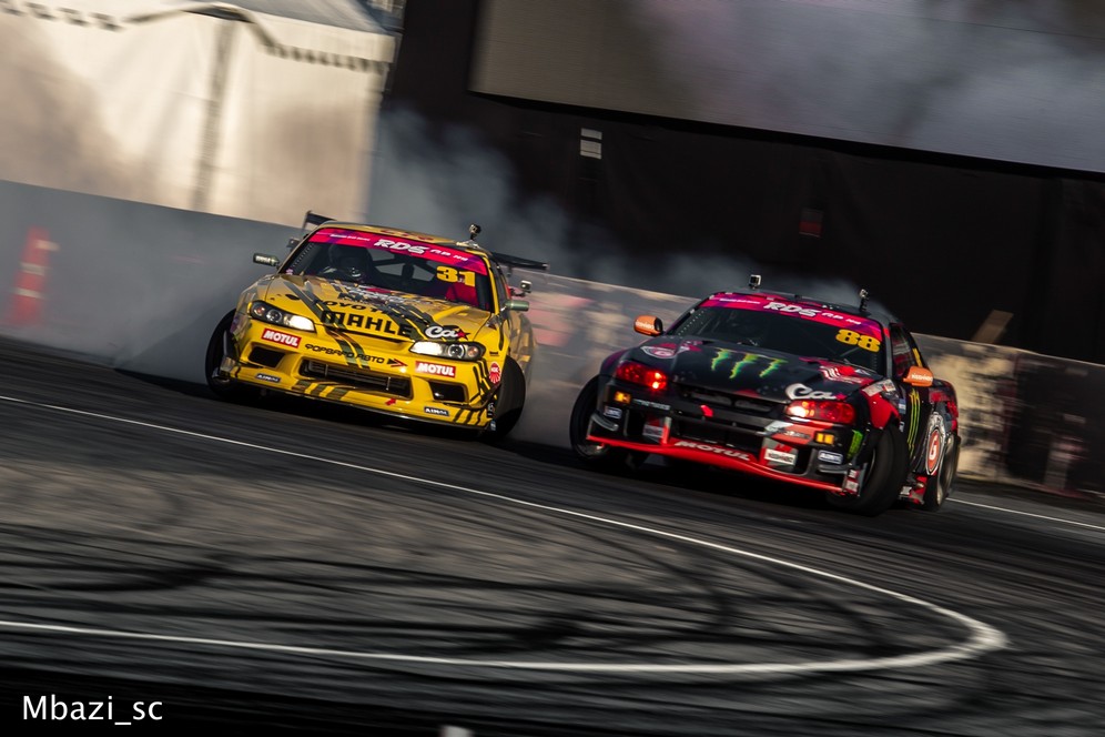 A yellow car and a black car skid round a bend in a stage of the Russian Drift Series car racing championship