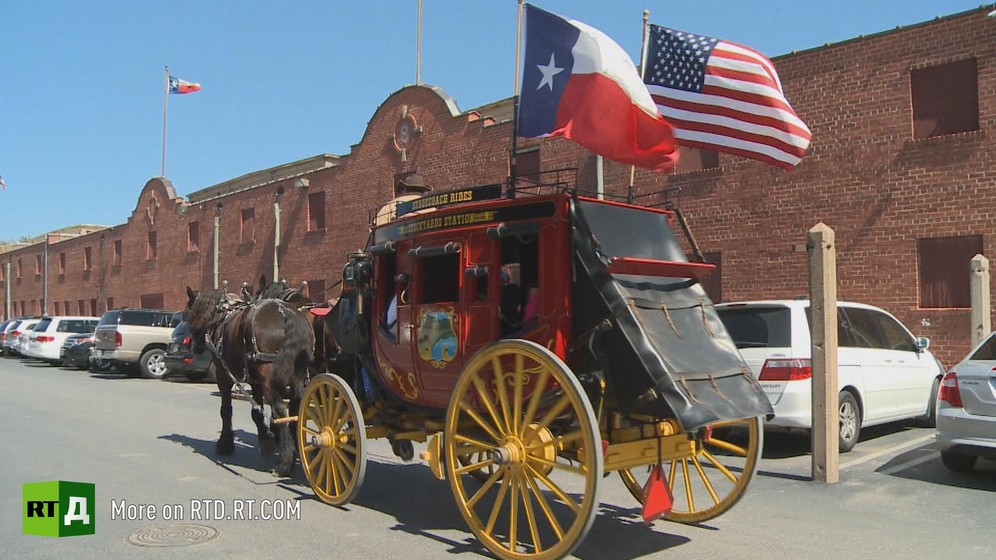 A red horsedrawn stagecoach with Texas and United States flags drives through a sunny street in Texas with low-rise red brick buildings in the background. Still taken from RTD documentary The REpublic of Texas.