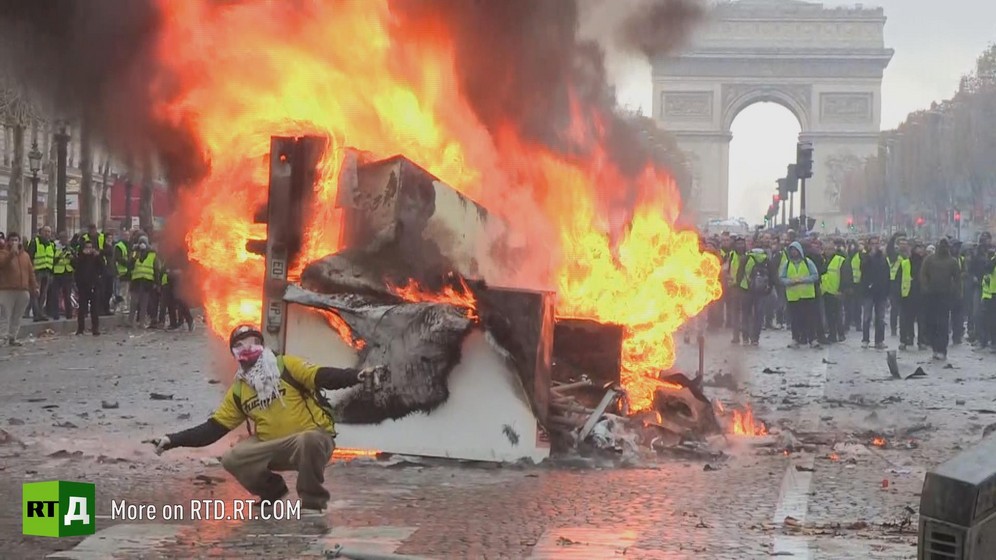 a Yellow Vest protestor wearing a mask  is crouching in front of a burning car, arms outstretched. In the background, a crowd of demonstrators wearing yellow vests are walking in front of the Arc de Triomphe in Paris. Still taken from RTD documentary Yellow Vest Fever.