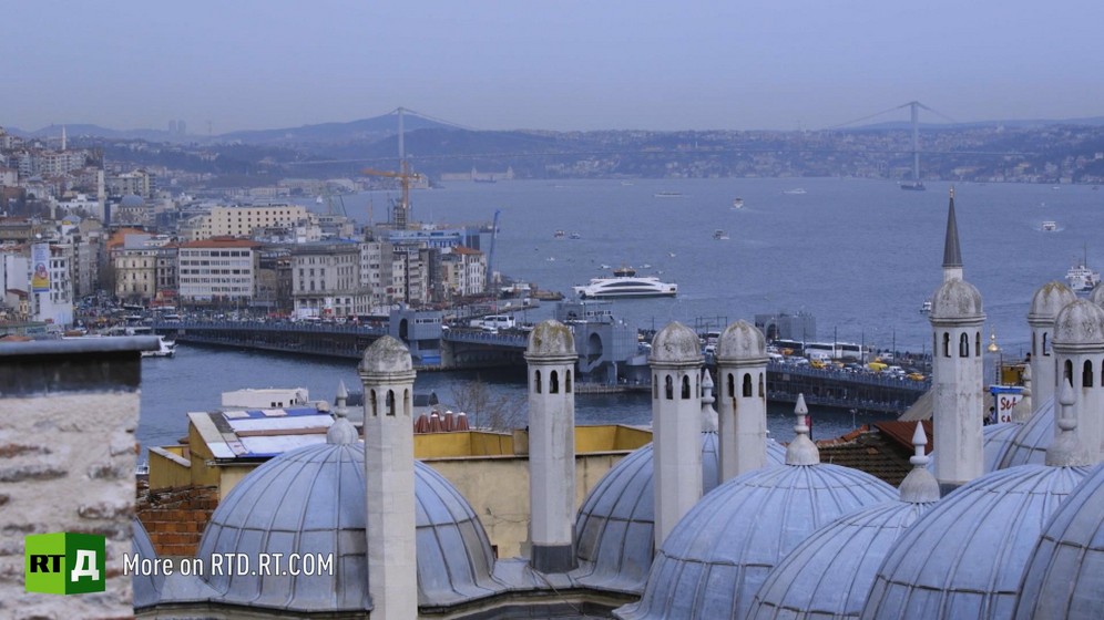 The Bosphorus in Istanbul, Turkey. In the foreground are the grey-blue domes and minaret of a mosque, then a ship next to the bank, with a bridge, the water and opposite bank of the straits in the background. Still taken from RTD's documentary series The Gulen Mystery.