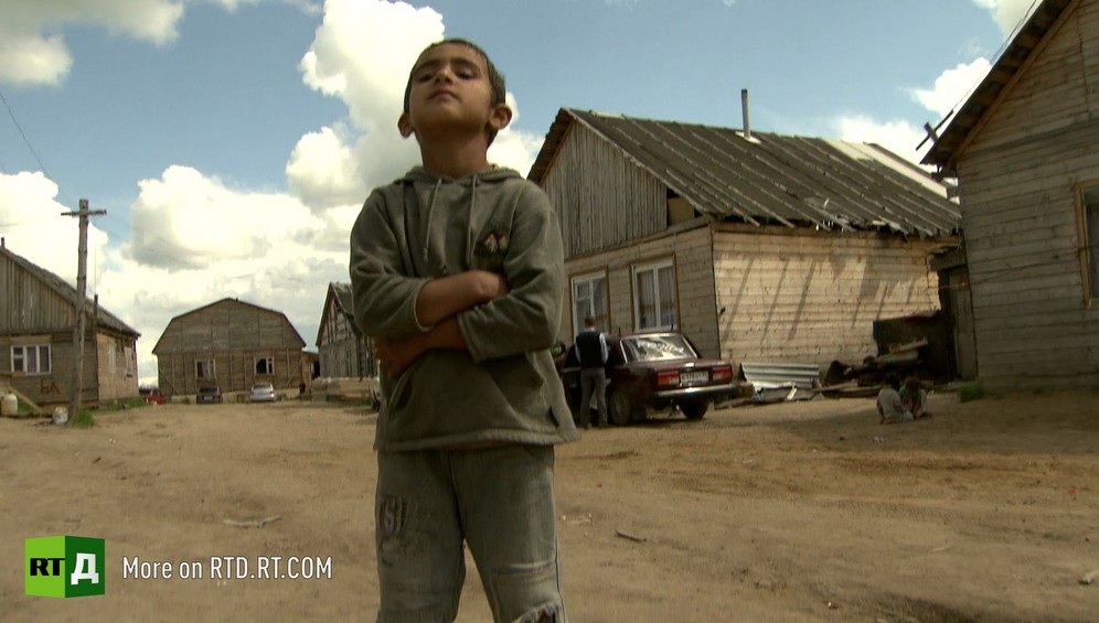 Boy stands with his nose in the air on a dirt road in front of wooden houses in a Kalderash Gypsy settlement in rural Russa.