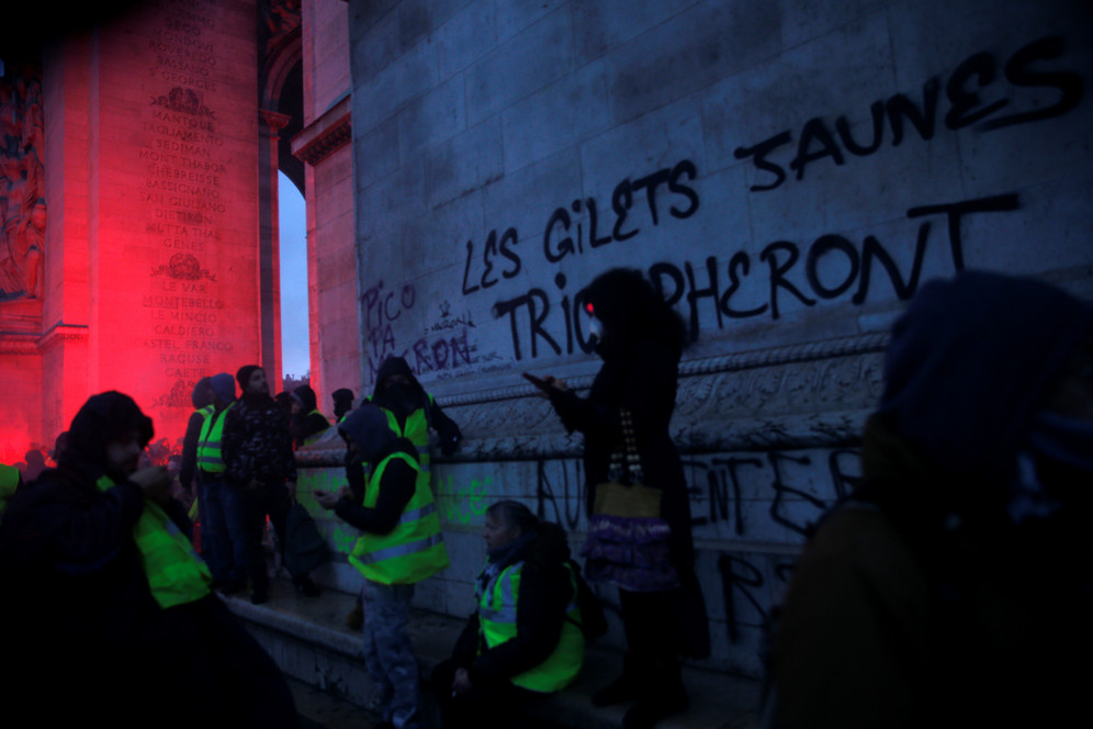 Masked protesters check their phones beneath the Arc de Triomphe painted with the slogan 