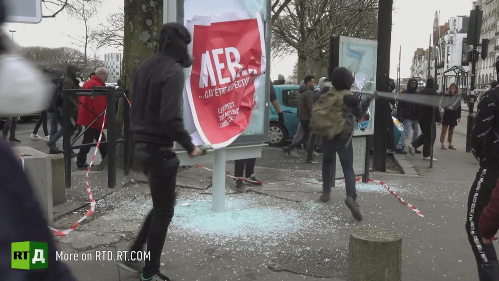 A person wearing a hoodie watches as another, also wearing a hoodie and a backpack, breaks the glass on urban furniture with an iron bar during a Yellow Vest protest in France.
Still taken from RTD 's documentary, Yellow Vest Fever.