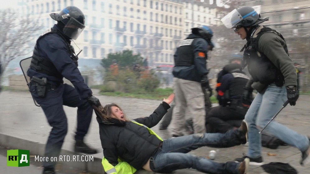One riot policeman wearing a helmet, carrying a shield and a baton, drags a female Yellow Vest protester by the hood along the ground, while another policeman in plain clothes and wearing a helmet with the visor up towers over her, with other policemen neutralising another protester in the background.
Still taken from RTD 's documentary, Yellow Vest Fever.
