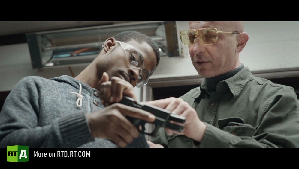 An African American teenage boy wearing safety glasses is being instructed to handle a gun by a middle aged White man as part of a Black Guns Matter training session. Still taken from RTD documentary Black Lives 5. Agents of Change