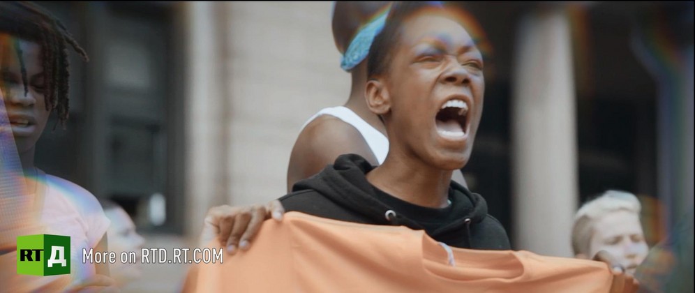 A Black boy wearing a Black hooded top is shouting while holding an orange sweatshirt, surrounded by protesters. Still taken from RTD documentary Black Lives 8: Deadlock.