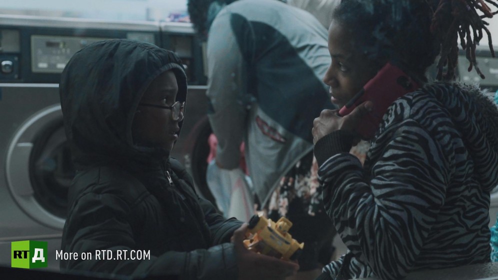 In a laundromat, an African American mother is speaking on her phone while her young son is holding a toy. Still taken from RTD documentary Black Lives 6. Illusion on single motherhood, teenage parenthood and the child poverty trap. 