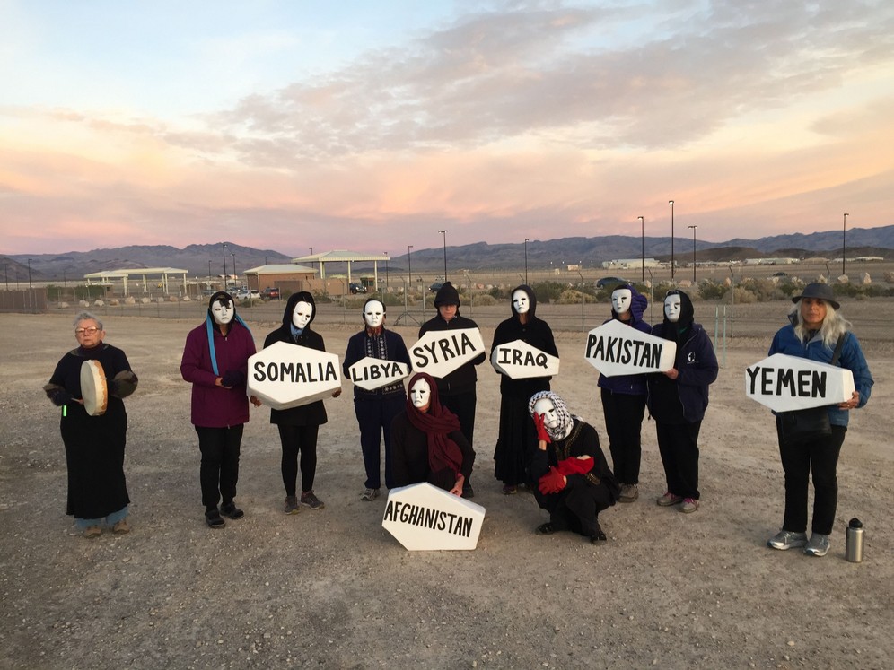 A group of women wearing white face maks are standing outside Creech Airbase in the Nevada desert, USA. They are holding small white coffins labelled 'Iraq", "Somalia", "Syria", "Pakistan", "Afghanistan", "Libya" and "Yemen". Taken while shooting RTD documentary on the impact of drones, Game of Drones.