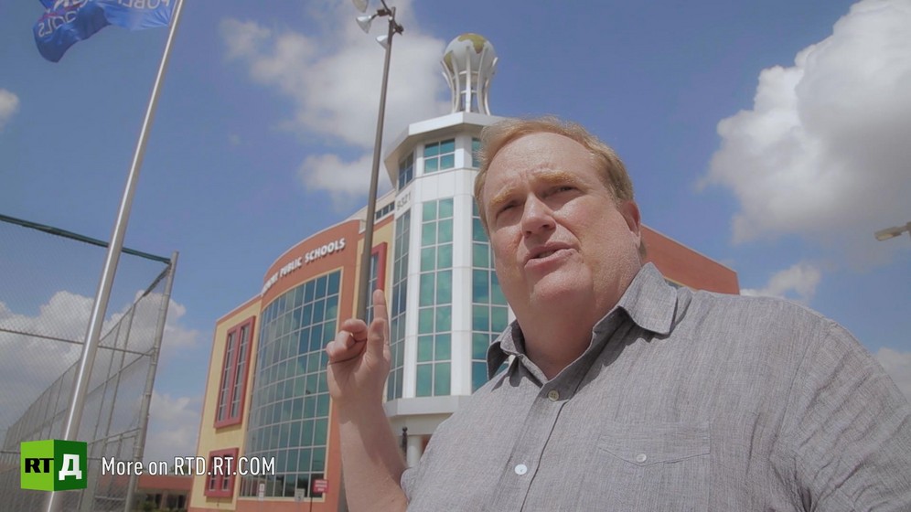 Mark Hall stands in front of a Harmony Public Schools building and points with his finger at the globe on top of one of its corners, in the United States. Still taken from RTD's documentary series on Fethullah Gulen, The Gulen Mystery, Episode 4: Gulen's Turkish Charter Schools.