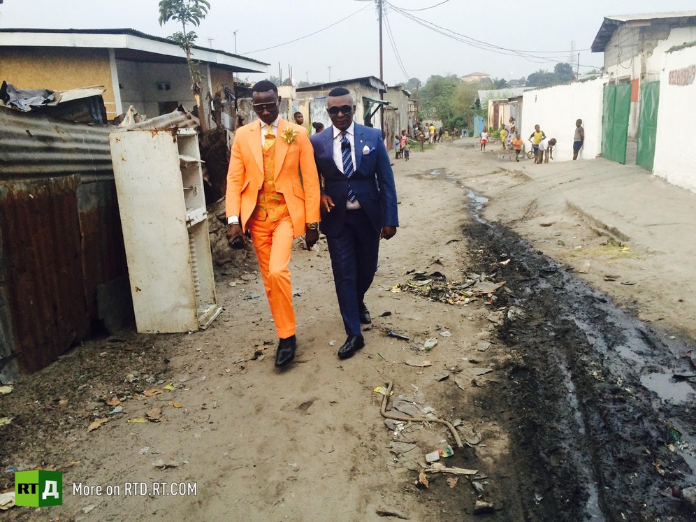 Maxime Dutoit wearing and orange suit walks with another Sapeur in the street in Brazzaville. Screenshot taken from RTD documentary Congo Dandies.