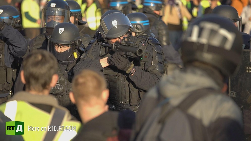 A Policeman in black wearing helmets aims a defense ball launcher while surrounded by police and Yellow Vest protesters.  Still taken from RTD 's documentary, Yellow Vest Fever.
